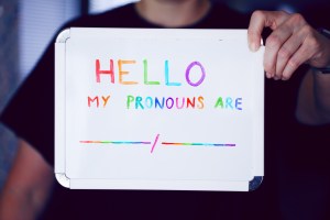 An image of a hand-held whiteboard displaying an introduction with pronouns included. The colours used are rainbow colours.